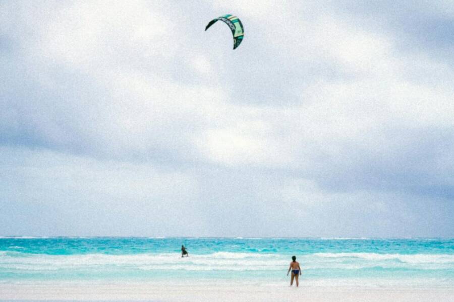 A person enjoying kitesurfing on a serene tropical beach with clear blue waters and cloudy skies.