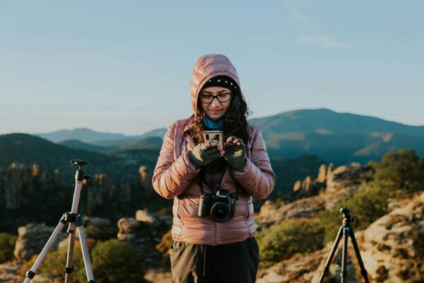 Female photographer in hooded jacket capturing stunning mountain scenery with a smartphone and tripod.