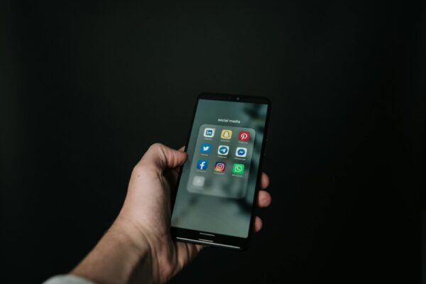 Close-up of a hand holding a smartphone displaying various social media app icons on a dark background.