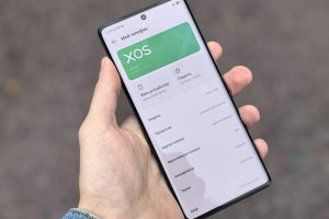 Person holding smartphone showing XOS interface on the screen, captured outdoors.