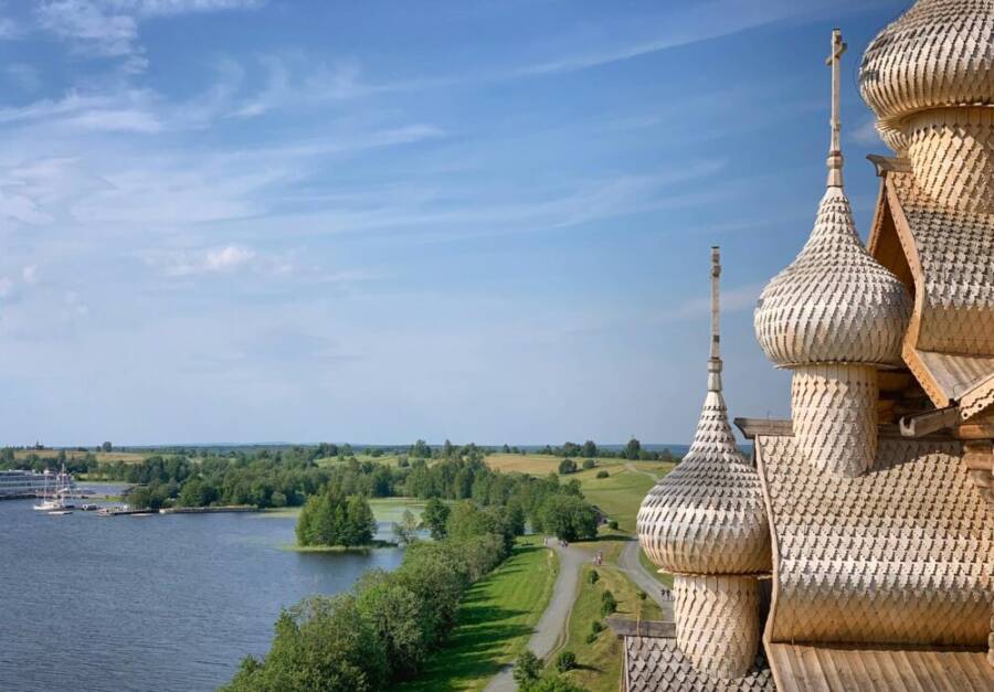 Wooden domes rise against a blue sky, overlooking a peaceful lake and lush greenery.