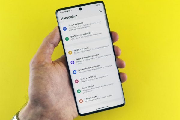 Close-up of a hand holding a smartphone with settings menu on a bright yellow background.
