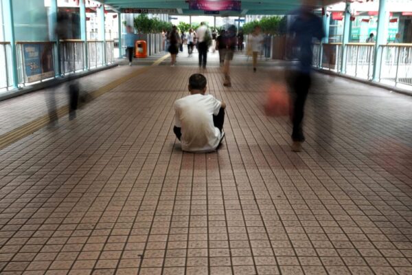 A solitary figure seated amidst blurred bustling pedestrians in a Hong Kong walkway.