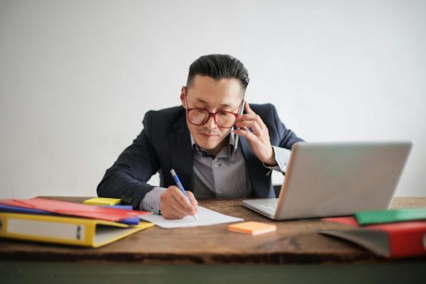 來電自動掛斷怎麼辦?封鎖與勿擾規則一步步教學 Professional businessman multitasking with phone call and laptop while writing notes at desk.