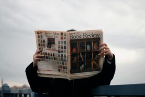 Individual holding a book review newspaper with colorful nails against a cloudy sky backdrop.