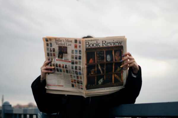 Individual holding a book review newspaper with colorful nails against a cloudy sky backdrop.