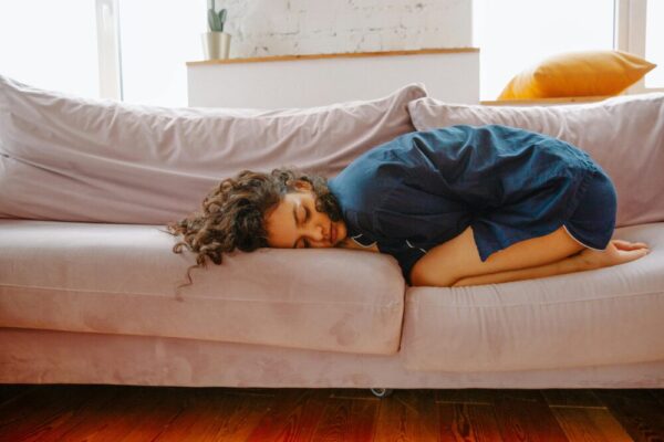 A woman rests on a sofa in pajamas, curled up in discomfort during the day.