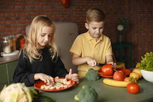 Focused little girl and boy cutting various vegetables and fruits while sitting at table and cooking in loft kitchen with brick wall