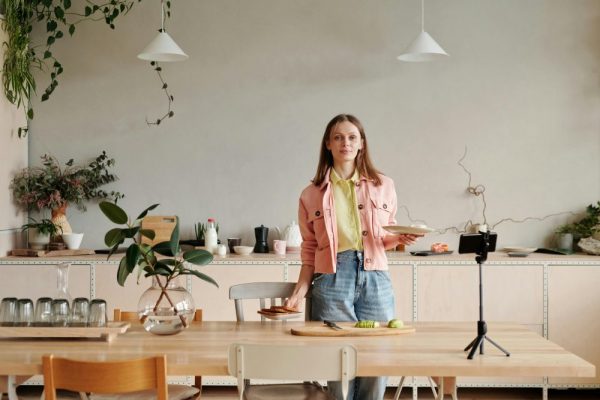 Young woman in kitchen setting up a cooking vlog with smartphone on tripod.