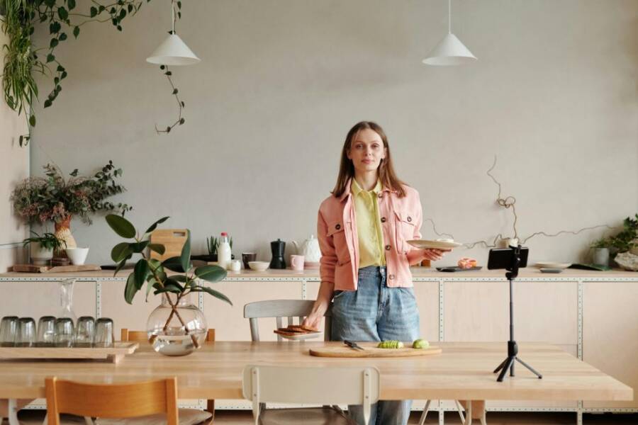Young woman in kitchen setting up a cooking vlog with smartphone on tripod.