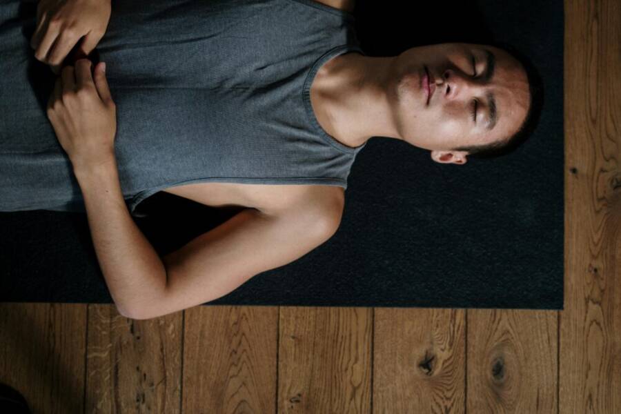 A man lies on his back in a yoga studio, relaxing on a mat in natural light.