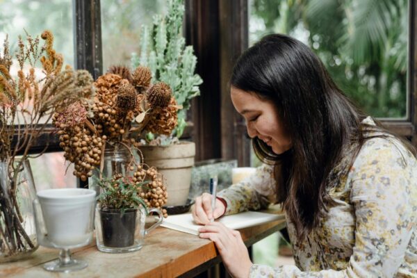 A cheerful young woman writes in a notebook surrounded by indoor plants, creating a cozy and creative workspace.