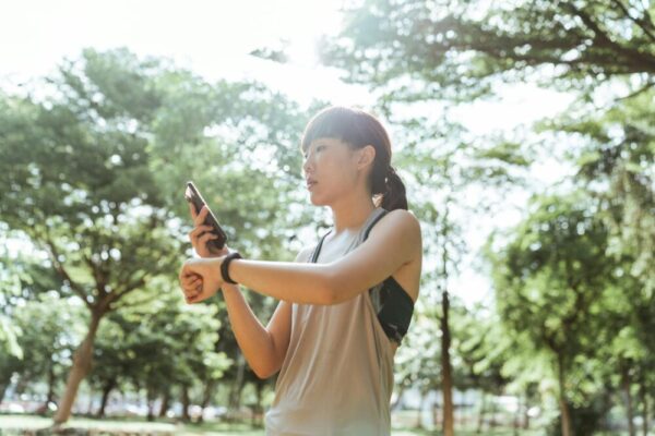 Low angle side view of fit Asian female athlete in wearable device and activewear surfing internet on cellphone during workout in park in sunlight in back lit