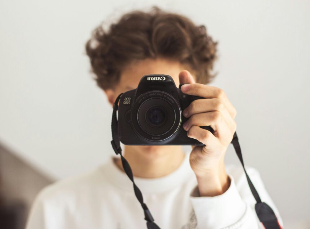 A young adult holding a Canon DSLR camera, capturing a portrait close-up.