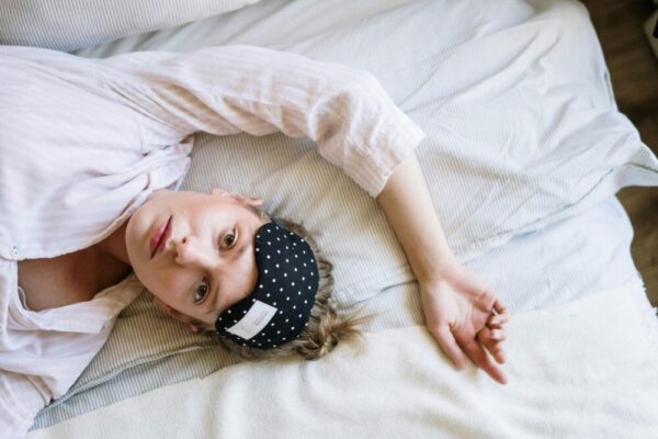 A young woman wears a polka dot eye mask while lying comfortably in bed during the day.