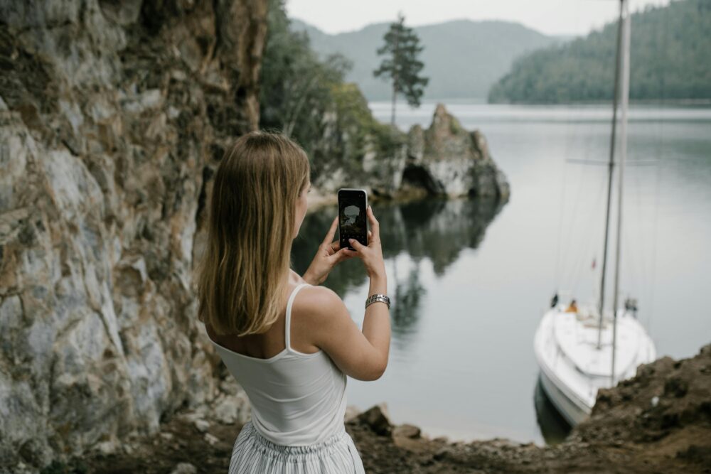 A woman takes a photo of a serene lakeside with a sailboat in lush natural surroundings.