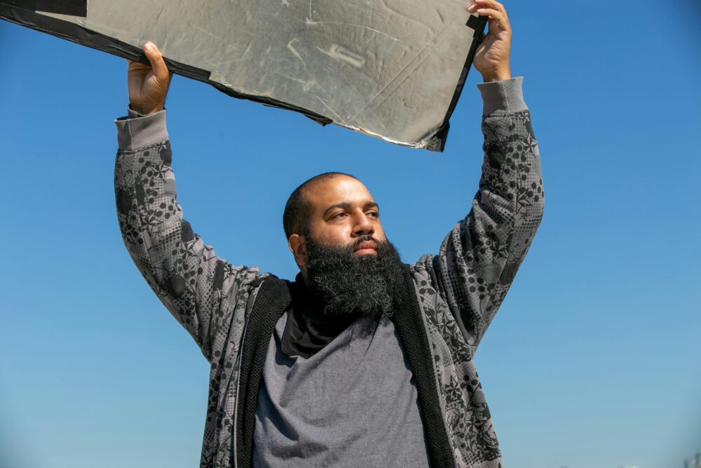 A man in casual clothing lifts a reflector under a clear blue sky, focusing sunlight.