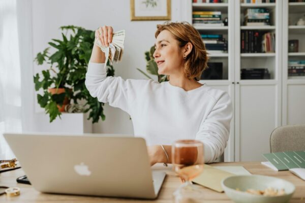 Smiling woman holding cash, working at home office with a laptop, symbolizing financial success.