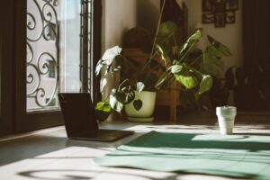 Inviting yoga setup with mat, laptop, and plants near a sunny window, promoting relaxation and focus indoors.