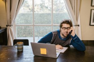 A man looking fatigued while working from home on his laptop with a coffee mug nearby.