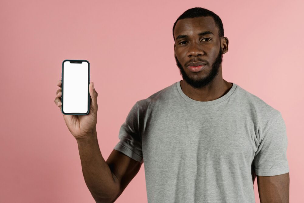 African American man presenting a blank smartphone screen against a pink backdrop.