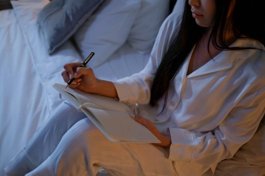 A woman in white pajamas writing in a journal while sitting on a bed. Cozy bedtime routine.