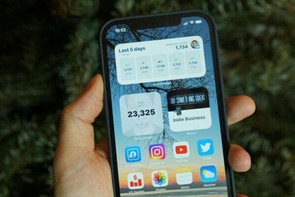 A hand holding a smartphone displaying various home screen widgets and app icons.