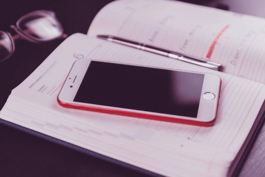 A red and white smartphone on an open diary accompanied by glasses and a pen.