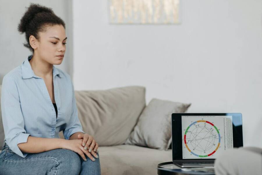 Young woman sitting in a living room analyzing an astrology chart on a laptop, reflecting concentration.