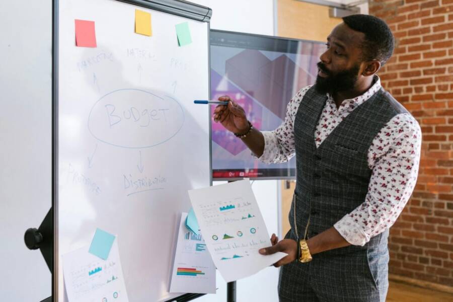 A businessman explains budget strategies using a whiteboard in an office setting.