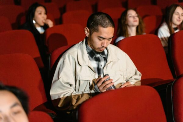 Asian man using phone in a movie theater while others watch the screen attentively.