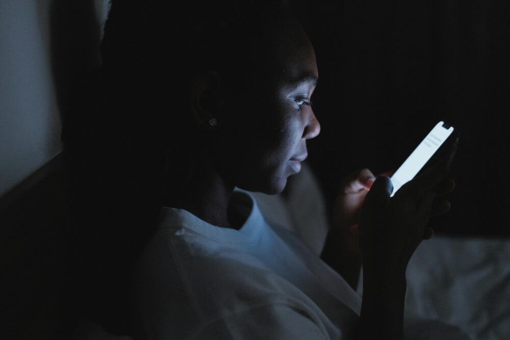 A woman using a smartphone in a dark bedroom, lit by the screen glow at night.
