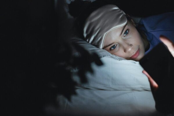 A woman in bed wearing a sleep mask uses her phone at night, creating a cozy ambiance
