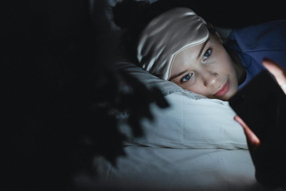A woman in bed wearing a sleep mask uses her phone at night, creating a cozy ambiance