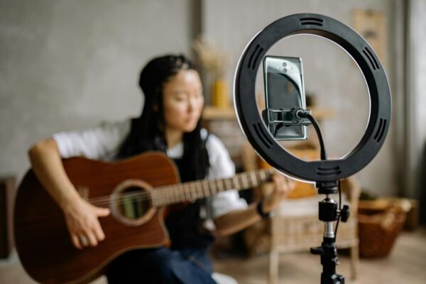Focused woman playing guitar during a live stream with a smartphone in a ring light indoors.