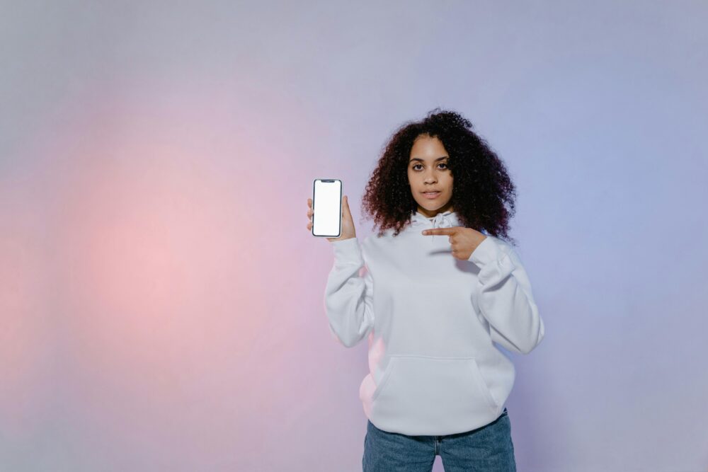 Woman in white hoodie pointing at a smartphone with blank screen, ideal for mockup designs.