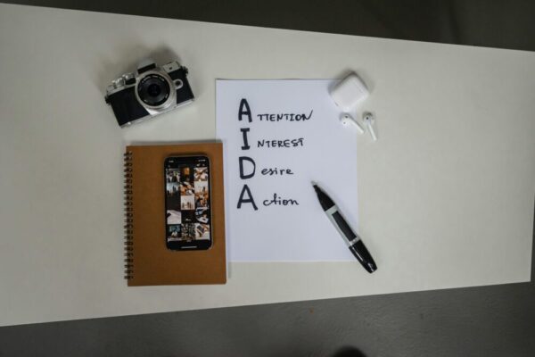 數位行銷 AIDA 模型是什麼?應用步驟、策略與實戰案例(含指標) Top view of AIDA marketing elements with notebook, smartphone, camera, and earbuds on a desk.