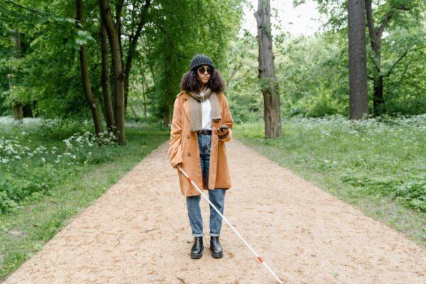 Stylish woman in coat explores a green park with a cane, using her smartphone for guidance.