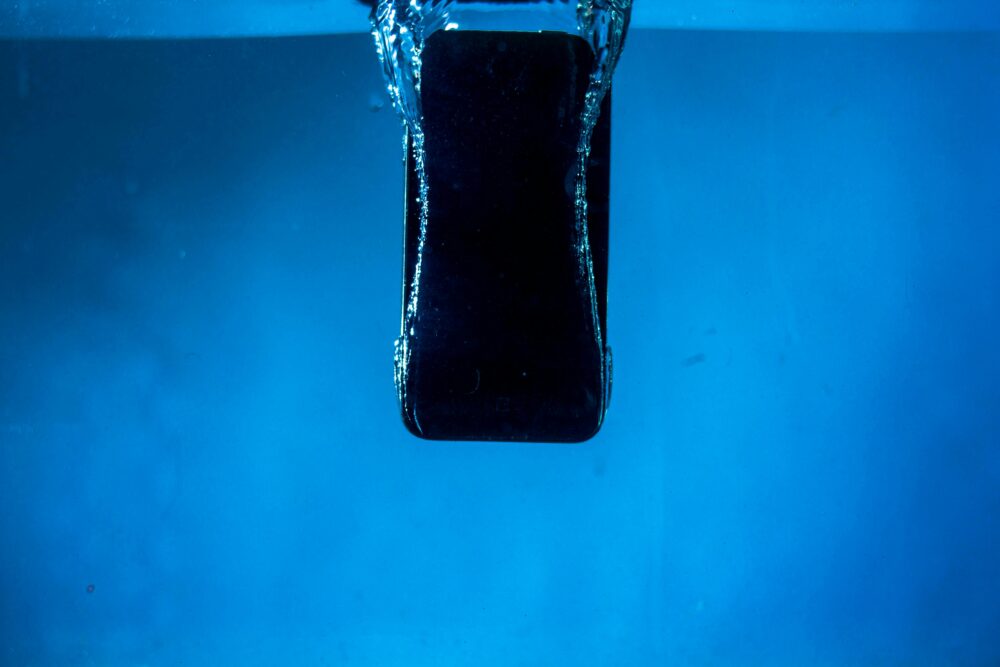 A smartphone partially submerged in water, captured against a blue backdrop.