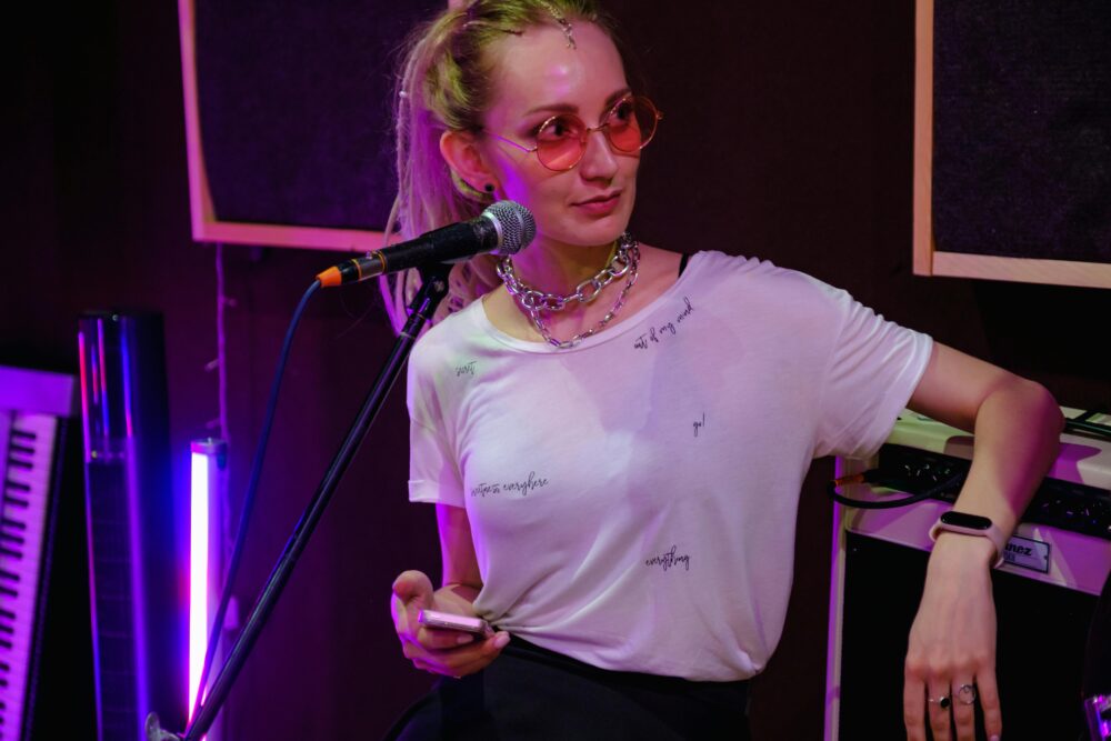 A woman musician in a studio holding a microphone and smartphone, wearing sunglasses and a chain necklace.