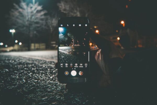 A hand holds a smartphone capturing a street scene at night with illuminated lights and bokeh.