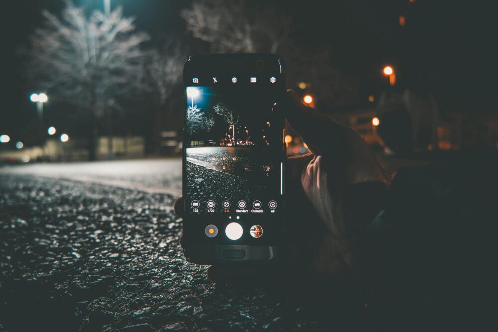 A hand holds a smartphone capturing a street scene at night with illuminated lights and bokeh.
