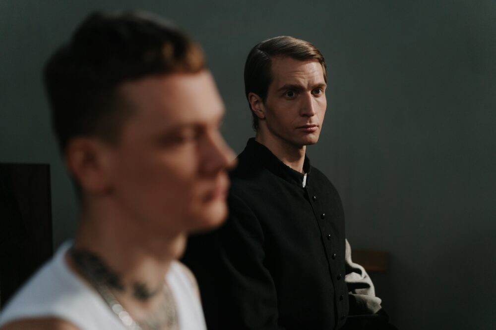 Two men, one in focus, sit thoughtfully indoors, highlighting contemplation.