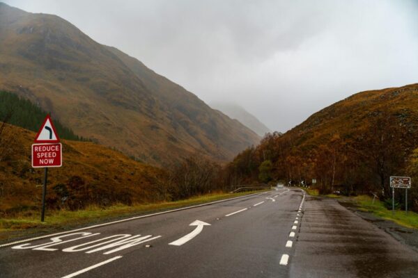 秋季的蘇格蘭高地，一條蜿蜒的道路，路邊設有樹木標誌，遠處的山脈籠罩在霧氣中。