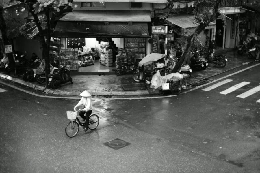 一張黑白照片，拍攝於越南雨中的街道，展現了一位騎自行車的人的都市生活。