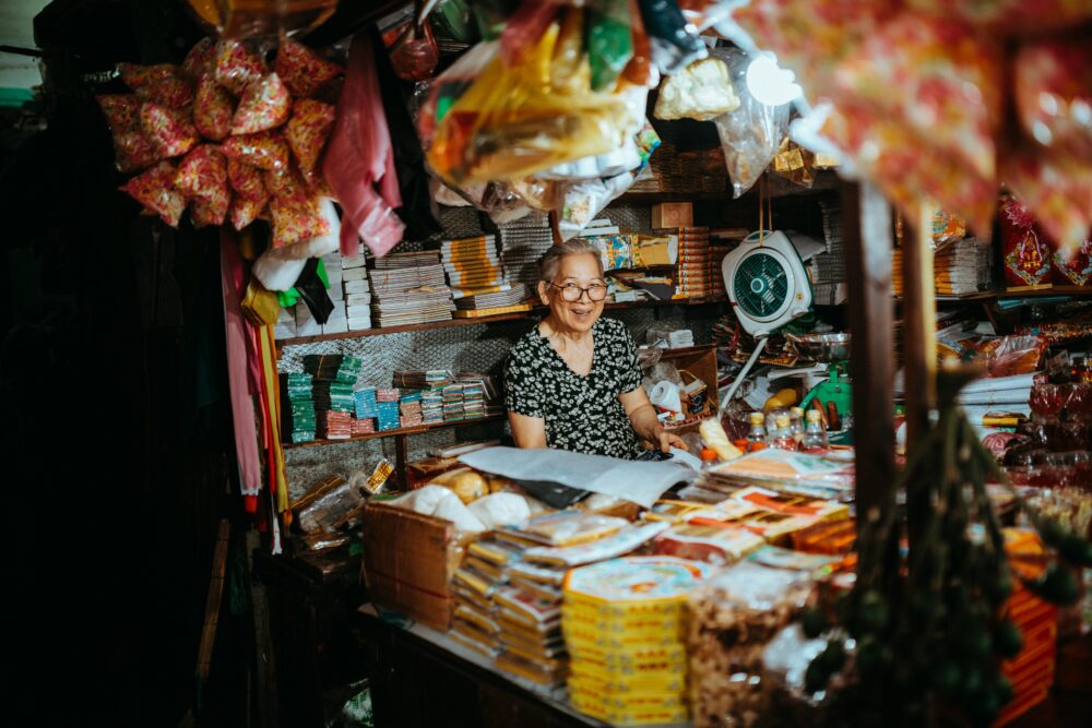 一位老婦人在越南市場攤位上擺滿了色彩繽紛的商品，微笑著。