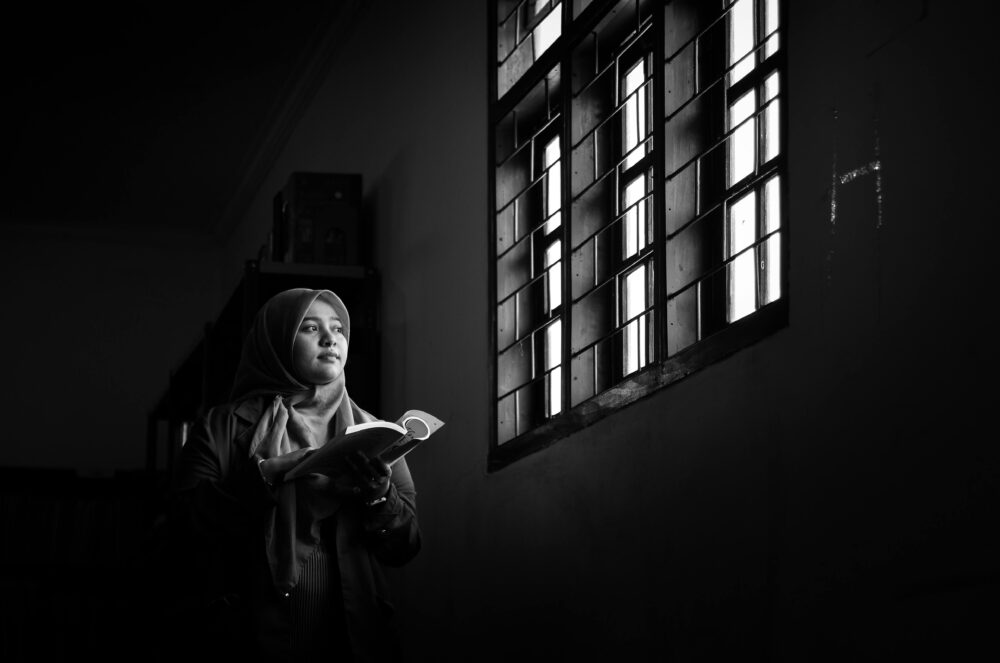 一張黑白照片,照片中一位女子在光線昏暗的房間裡靠窗讀書,引人深思。