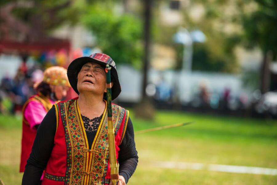 An elderly Taiwanese woman in vibrant traditional attire participates in an outdoor cultural event.