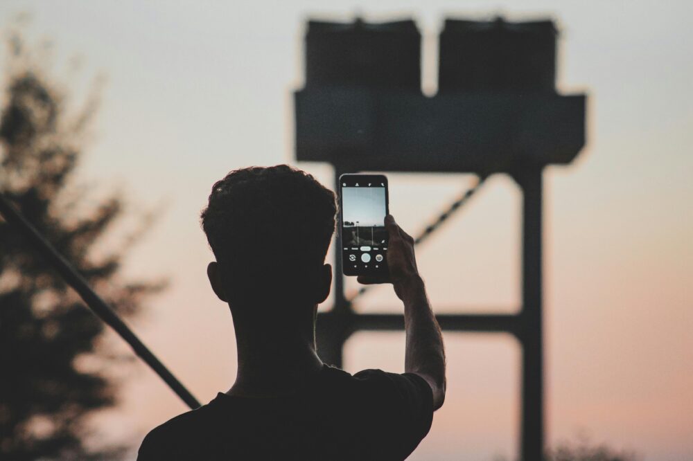 A man silhouettes against a sunset, capturing the view with a smartphone outdoors