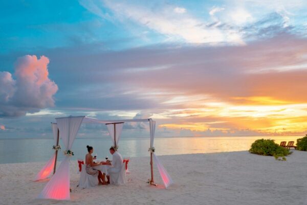 A couple enjoys a romantic beach dinner under a canopy at sunset, creating a serene and scenic atmosphere.